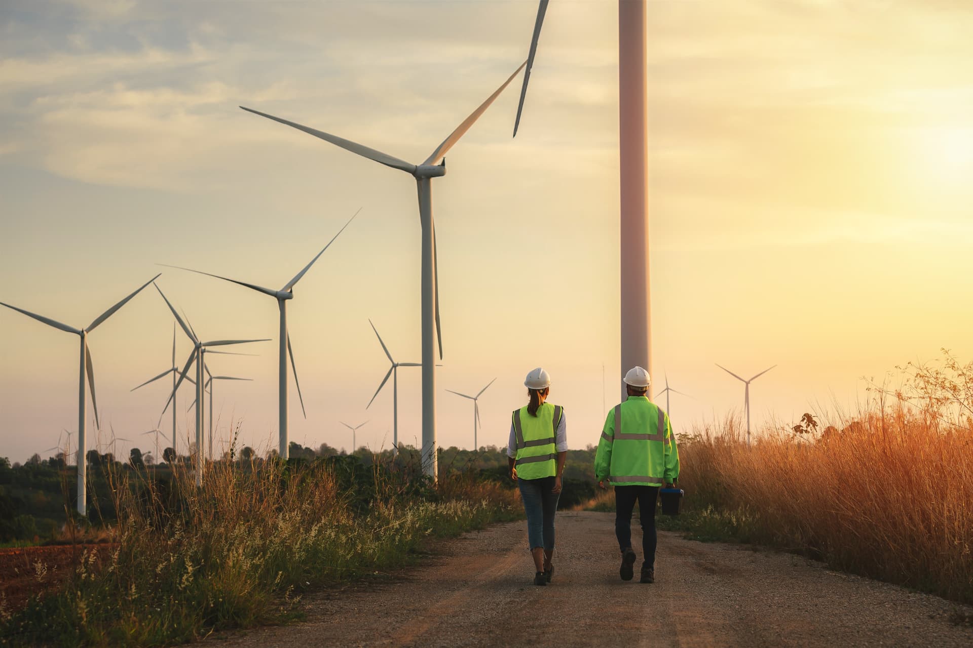 Two technicians in high-visibility vests walking a track through a wind farm at sunset
